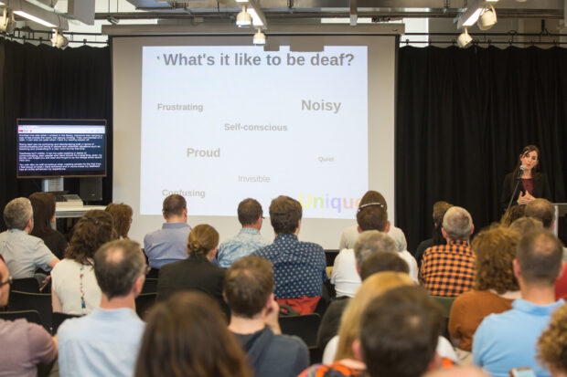 An audience listening to a talk with a slide saying 'what it's like to be deaf' in the background