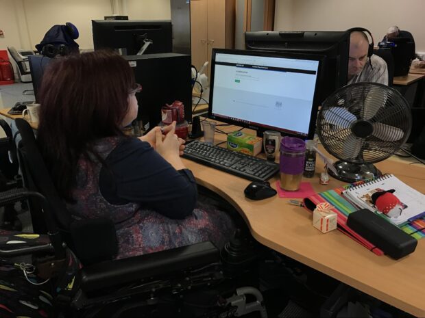 A photo of Becs sat in front of a computer. She is sat in a wheelchair with her hands clasped together.