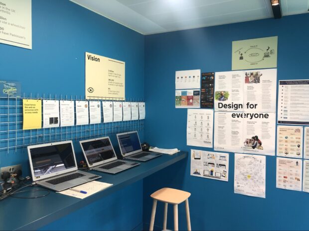 Three Chrome laptops on a long desk with a stool underneath, posters on two surrounding with headings of Vision and Design for Everyone.