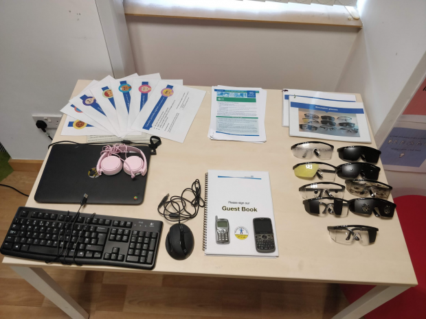 A table with a laptop, headphones, keyboard, mouse, 9 pairs of simulation glasses, 2 phones and reading material
