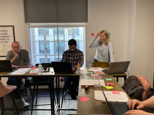 The Delivery Manager and 1 developer sat at a high table in a meeting room testing the service using the accessibility personas with the Designer standing behind observing.